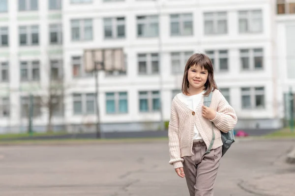 Menina feliz uma estudante de 8 anos de aparência europeia com uma mochila está andando no quintal da escola à tarde na rua olhando para o close-up da câmera — Fotografia de Stock