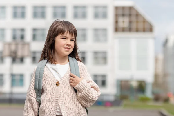 Chica feliz una colegiala de 8 años de apariencia europea con una mochila está caminando en el patio de la escuela por la tarde en la calle mirando la cámara de cerca —  Fotos de Stock