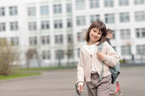 Chica feliz una colegiala de 8 años de apariencia europea con una mochila está caminando en el patio de la escuela por la tarde en la calle mirando la cámara de cerca —  Fotos de Stock