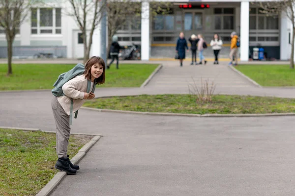 Ragazza felice una studentessa di 8 anni di aspetto europeo con uno zaino sta camminando nel cortile della scuola nel pomeriggio per strada guardando la fotocamera primo piano — Foto Stock