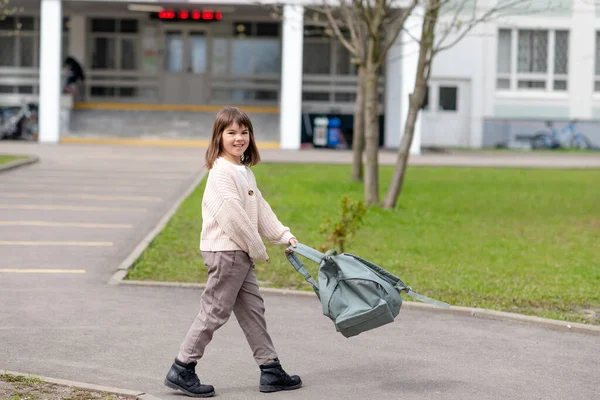 Chica feliz una colegiala de 8 años de apariencia europea con una mochila está caminando en el patio de la escuela por la tarde en la calle mirando la cámara de cerca —  Fotos de Stock