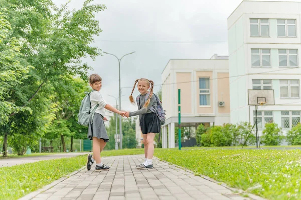 Duas lindas meninas felizes alunas com mochilas estão jogando perto da escola — Fotografia de Stock