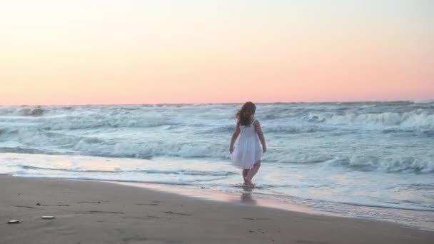 Kleines Mädchen in langer Kleidung mit langen Haaren läuft im Sommer bei Sonnenuntergang am Strand entlang — Stockvideo