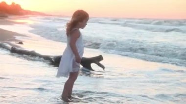 Yazın gün batımında deniz kıyısında uzun saçlı küçük bir kız çocuğu kumsalda koşuyor.