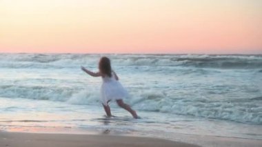 Yazın gün batımında deniz kıyısında uzun saçlı küçük bir kız çocuğu kumsalda koşuyor.