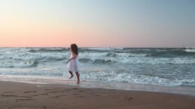 Yazın gün batımında deniz kıyısında uzun saçlı küçük bir kız çocuğu kumsalda koşuyor.