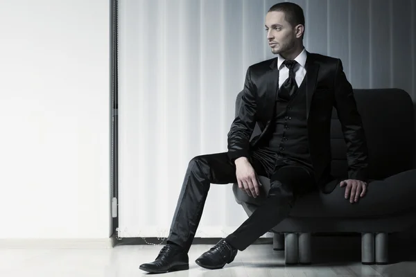 Elegant young fashion man in tuxedo on a sofa, blinds background — Stock Photo, Image