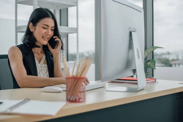 Genç Asyalı kadın dizüstü bilgisayar kullanıyor ve telefonla konuşuyor..