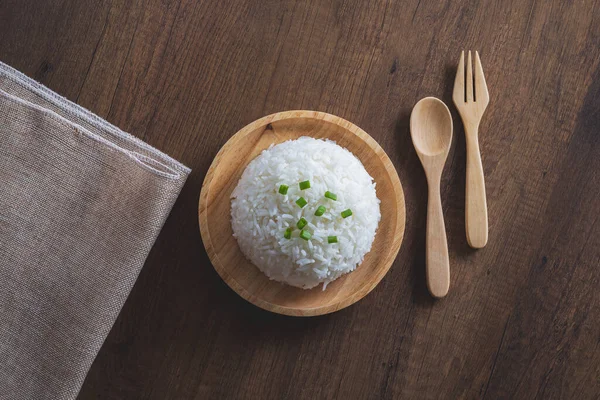 Cooked white rice in a wooden dish with green onion sliced on top with wooden spoon, wooden fork and placemat on a wooden table. Thai Jasmine rice.