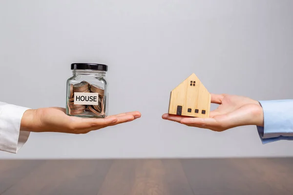 Client hand holding a money jar and house model in home broker hand or salesman person. on wooden desk office.