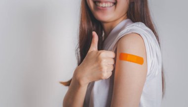 Asian woman showing thumb up gesture and shows off an orange bandage after receiving the covid-19 vaccine. Vaccination campaign concept for safe life return to normal life with copy space for text.