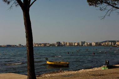 Vlor, Arnavutluk 17 Ağustos 2025: İnsanlar Vlora 'nın sıcak kıyı sularında yüzerken kıyı şeridi yakınlarında küçük sarı bir tekne duruyor.