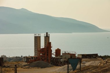 Arnavutluk 'un Vlora kenti yakınlarındaki denize bakan sanayi şantiyesi 17.08.2025' te fotoğraflandı. Makineler ve depolama siloları sakin bir su zeminine ve katmanlı dağ siluetlerine karşı dururlar, sıcak akşam ışığında yıkanırlar..