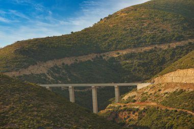 Vlor, Arnavutluk 17 Ağustos 2025: Engebeli yeşil tepeler boyunca uzanan yüksek beton bir köprünün uzak bir görüntüsü, sıcak akşam ışıklarıyla aydınlanıyor.