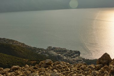 Vlor, Arnavutluk 17 Ağustos 2025: Işıldayan denizin üzerindeki uzak dağlar yükselirken, önplanda kurak yaz bitkilerini aydınlatan yumuşak altın ışıklı sakin bir kıyı manzarası. Sisli gökyüzü ve yumuşak güneş ışığı bir sakinlik yaratır..