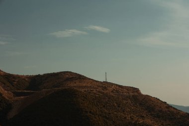 Vlor, Arnavutluk 17 Ağustos 2025: Dağılmış bitki örtüsüyle kaplı ve uzun elektrik direkleriyle ve elektrik hatlarıyla kesişen, sıcak altın tonlarıyla engebeli bir dağ manzarası