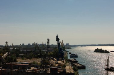 Açık bir günde uzak bir şehrin siluetine karşı büyük vinçleri, taşıyıcı yapıları, yanaşmış gemileri ve depoları olan bir sanayi limanı manzarası..