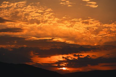 Karanlık dağ siluetlerinin üzerinde turuncu ve kırmızı tonlarda parlayan katmanlı bulutlarla dramatik, ateşli günbatımı, Ağustos 2025 'te yakalandı.