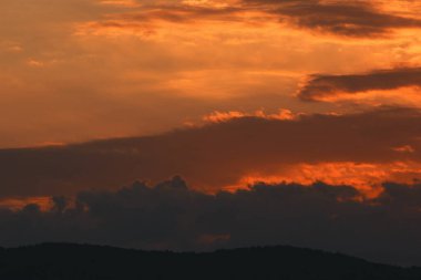 Karanlık dağ siluetlerinin üzerinde turuncu ve kırmızı tonlarda parlayan katmanlı bulutlarla dramatik, ateşli günbatımı, Ağustos 2025 'te yakalandı.