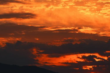 Karanlık dağ siluetlerinin üzerinde turuncu ve kırmızı tonlarda parlayan katmanlı bulutlarla dramatik, ateşli günbatımı, Ağustos 2025 'te yakalandı.