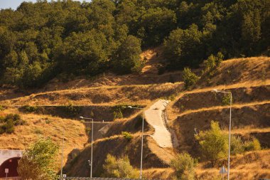Kuru altın çimleri ve yeşil ağaçlarla kaplı güneşli bir yamaçta dolambaçlı bir toprak yol ve yol kenarı altyapısı yükseliyor..
