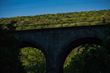 Ağustos 2025 'te, yoğun yaz yaprakları ve açık mavi gökyüzü ile birlikte, yeşil orman vadisinden geçen taş kemer köprüsü, doğal bir manzara içindeki tarihi altyapıyı vurguluyor..