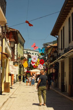 Vlor, Arnavutluk 20 Ağustos 2025: Yoğun yaz güneşinin altında, renkli sokak dekorasyonlarını, ahşap panjurlarla geleneksel mimariyi ve dar alanda yürüyen turistleri gösteren, tarihi eski kentte canlı bir gün ortası sahnesi.