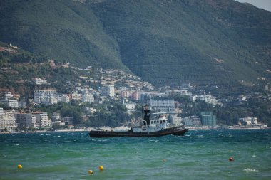 Vlor, Arnavutluk 21 Ağustos 2025: Bir römorkör kıyı kenti ve arka planda yükselen yemyeşil Vlor dağları ile canlı mavi denizi geçiyor.