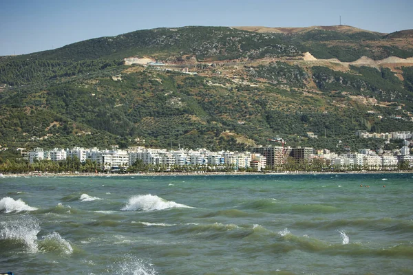 Arnavutluk 'un Vlora kentinde Ağustos 2025' te yakalanan sakin bir kıyı manzarası. Ufukta yükselen uzak tepeler gibi yumuşak gün ışığında hafif dalgalar parıldıyor. Birkaç kuş sakince gökyüzünde süzülerek huzurlu deniz manzarasına doğal bir aksan katıyor..