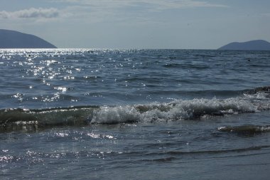 Arnavutluk 'un Vlora kenti Ağustos 2025' te kıyı kenti ve arka planda görünen yeşil Vlora dağlarıyla birlikte kıyıya doğru yumuşak dalgalar akmaktadır. Sahne, açık bir yaz gününde denizin sakin hareketini yakalar..