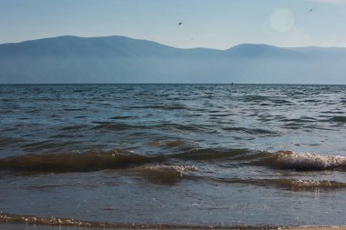 Arnavutluk 'un Vlora kentinde Ağustos 2025' te yakalanan sisli dağ siluetlerine karşı kıyıya yumuşak dalgalar vuruyor. Yumuşak ışık ve doğal dokularla sakin bir sahil manzarası..