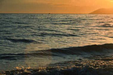 Altın gün batımı ışığı, 24 Ağustos 2025 'te Arnavutluk' un Vlora kentindeki yumuşak deniz dalgalarını yansıtarak yumuşak dalgalar ve uzak dağ siluetleriyle sıcak ve sakin bir kıyı atmosferi yaratıyor..