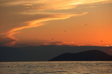 Altın gün batımı ışığı, 24 Ağustos 2025 'te Arnavutluk' un Vlora kentindeki yumuşak deniz dalgalarını yansıtarak yumuşak dalgalar ve uzak dağ siluetleriyle sıcak ve sakin bir kıyı atmosferi yaratıyor..