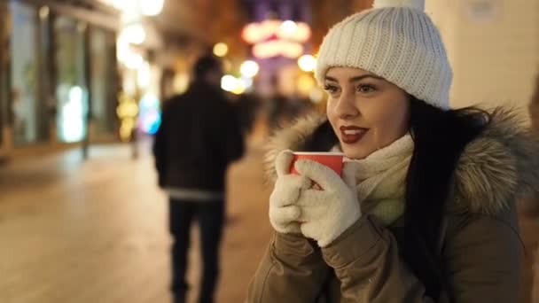 若い美人女性がスローモーションHdビデオで路上で寒い冬の夜にマルチワインを飲んでいます 彼女は笑っている 背景にぼやけたクリスマスライトと歩行者を光る — ストック動画