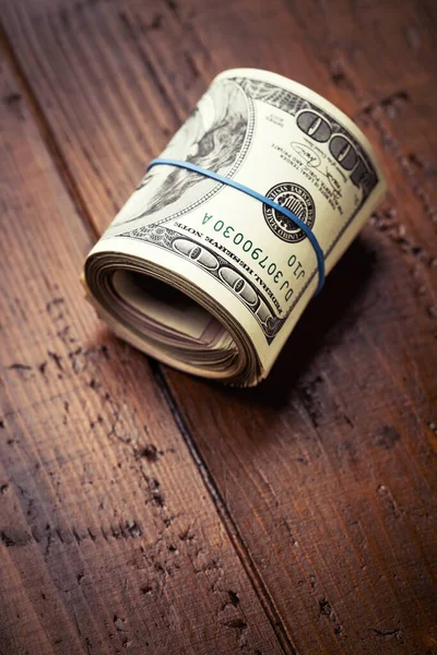 Roll of money with rubber on wooden table - one hundred US banknotes with president Franklin portrait. Cash of hundred dollar bills, paper currency background.