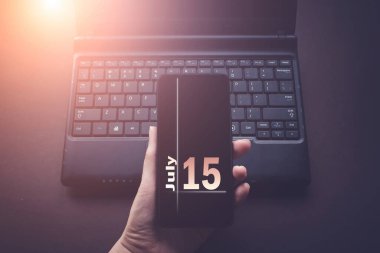 15 Temmuz. Ayın 15 'i, takvim tarihi. Laptop bilgisayarındaki el bilgisayarı, güneş ışığı alan karanlık bir arka plan. Yaz ayı, yılın günü konsepti.