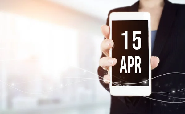 April 15th. Day 15 of month, Calendar date. White smartphone with Calendar date in businesswoman hand on blurred background. Spring month, day of the year concept