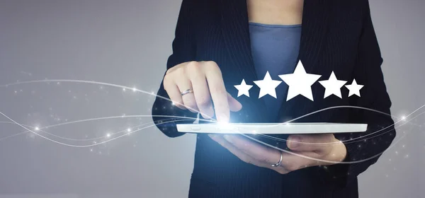 Hand pointing five star symbol to increase rating of company. White tablet in businesswoman hand with digital hologram Five stars 5 rating sign on grey background