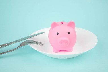 Spending Savings concept, piggy bank on white plate with fork and knife on blue background.