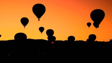 Gün batımında tepenin üzerinden uçan bir grup sıcak hava balonu silueti.
