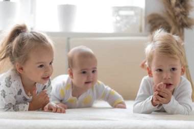 Üç küçük gülümseyen çocuk evdeki yumuşak beyaz yatakta birlikte oynuyorlar. Küçük yaşta bebekleri olan iki kardeş. Çocuklar yeni doğan kardeşlerle tanışıyor. Yeni yürümeye başlayan çocuklar gülüyor ve kaynaşıyor. Komik. Uzayı kopyala