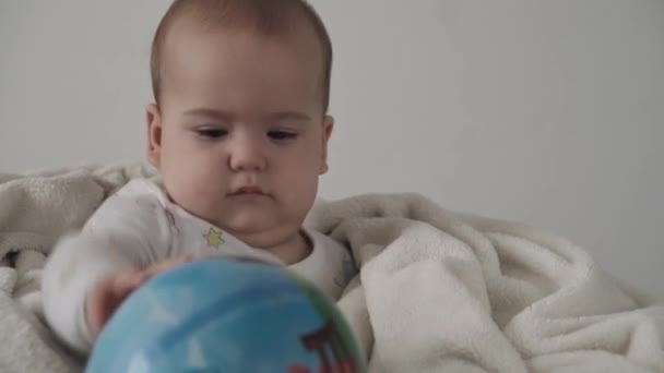 Bébé, concept d'enfance - gros plan de mignon sourire heureux drôle brun aux yeux potelés enfant nouveau-né éveillé. édenté 7 mois bébé jouer avec ballon assis sur lit blanc doux enveloppé dans une couverture chaude à la maison — Video