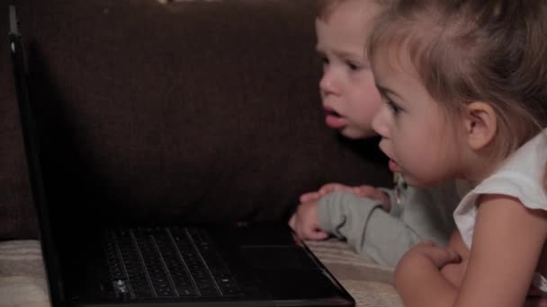 Família, jogo, quarentena, conceitos de infância - close-up Duas crianças felizes assistindo desenhos animados no laptop deitado no sofá marrom em casa durante a quarentena. Crianças irmãos irmão, irmã se divertir — Vídeo de Stock