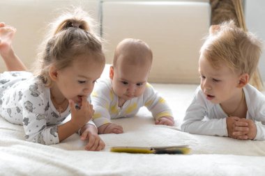 Üç küçük gülümseyen çocuk evdeki yumuşak beyaz yatakta birlikte oynuyorlar. Kardeşim ve kız kardeşim bebekli masal okuyorlar. Çocuklar yeni doğan kardeşlerle tanışıyor. Yeni yürümeye başlayan çocuklar gülüyor ve kaynaşıyor. Komik. Uzayı kopyala