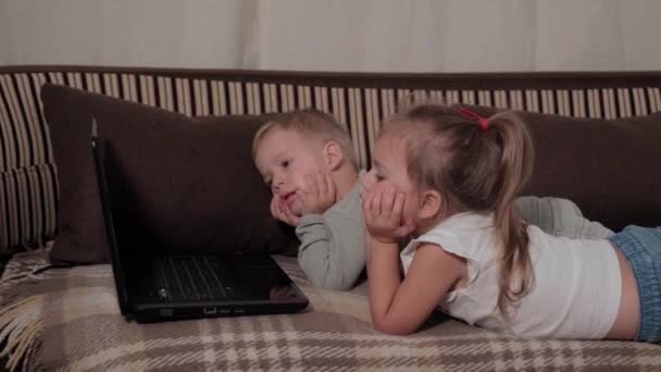Familie, spel, quarantaine, jeugdconcepten - Twee gelukkige jonge kinderen die tekenfilms kijken op een laptop die thuis op een bruine bank ligt tijdens de quarantaine. Kinderen broers en zussen broer, zus veel plezier samen — Stockvideo