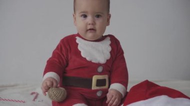 Mutlu Noeller, mutlu yıllar, bebekler, çocukluk, tatil konsepti - yakın plan gülümseyen 7 aylık Noel Baba şapkalı komik bebek, kırmızı tulum sıcak yumuşak yatağın üzerinde oturuyor tatlı Noel şekerini yalıyor
