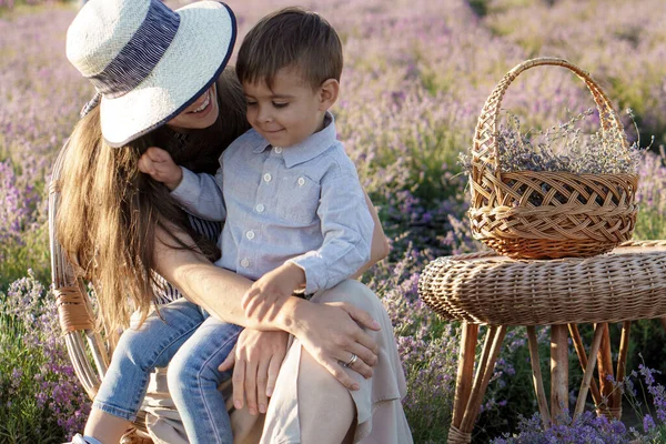 Keluarga besar, kebapakan, masa kecil, keibuan, konsep gaya provence - ibu muda duduk di kursi wicker dengan anak laki-laki kecil anak laki-laki dengan keranjang dan meja di lapangan lavender pada musim panas sebelum matahari terbenam — Stok Foto