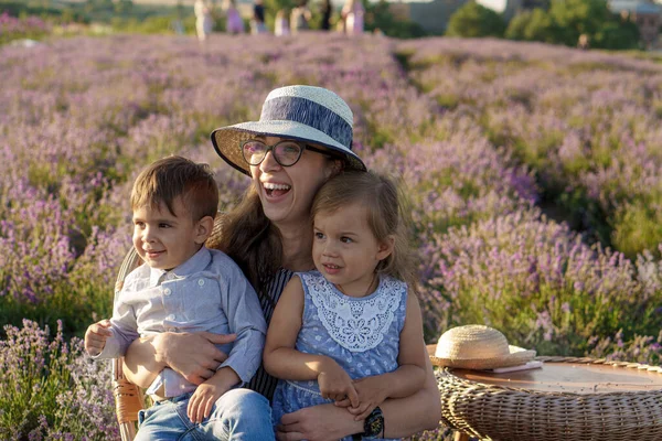 Grande família, paternidade, infância, maternidade, conceito de estilo de provence - a jovem mãe senta-se na cadeira de vime com o menino menino e a filha no campo de lavanda na noite de verão antes do pôr do sol — Fotografia de Stock