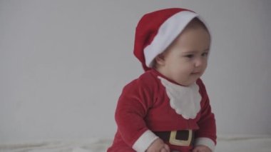 Mutlu Noeller, mutlu yıllar, bebekler, çocukluk, tatil konsepti - yakın plan gülümseyen 7 aylık Noel Baba şapkalı komik bebek, kırmızı tulum sıcak yumuşak yatağın üzerinde oturuyor tatlı Noel şekerini yalıyor