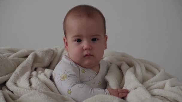 Criança, conceito de infância - close-up de sorrir feliz rosto gordinho engraçado de olhos castanhos de criança recém-nascida acordada. toothless 7 meses bebê grimaces, fecha os olhos sentar-se em cama macia envolto em cobertor quente em casa — Vídeo de Stock